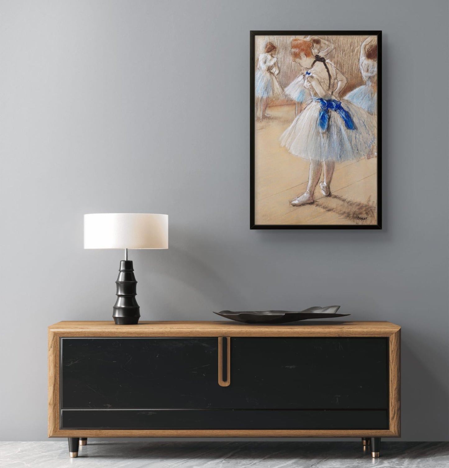 Framed artwork of a dancer with a lamp and decorative plate on a wooden cabinet against a gray wall.