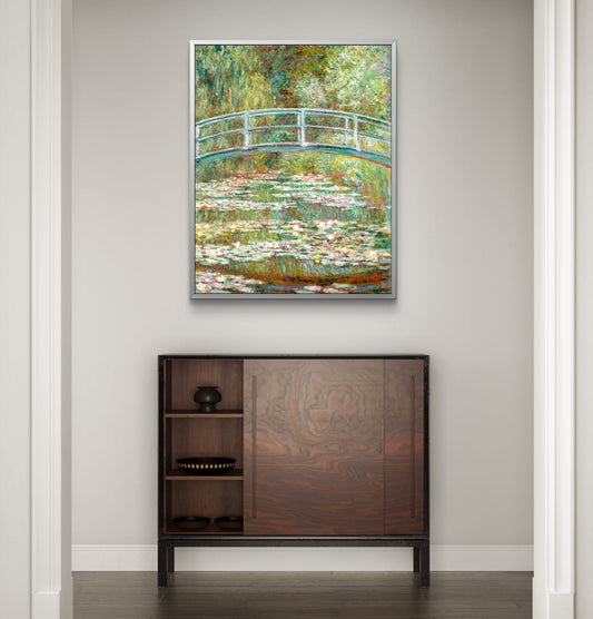 Framed painting of a bridge over water lilies above a wooden cabinet.